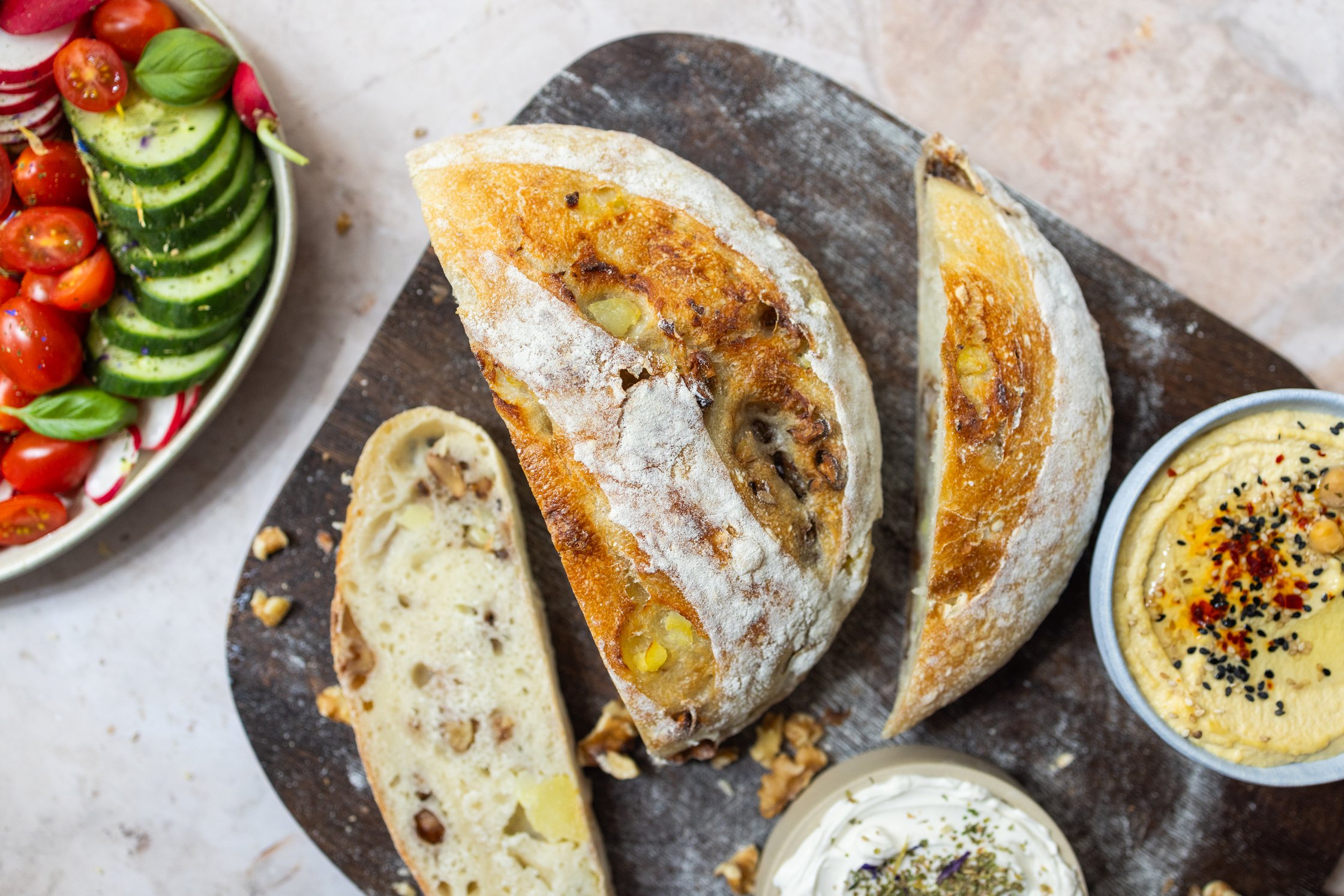 Geschnittenes Sauerteig brot auf einem Brett mit frischem Gemüse, Hummus und veganem Frischkäse in Schüsseln