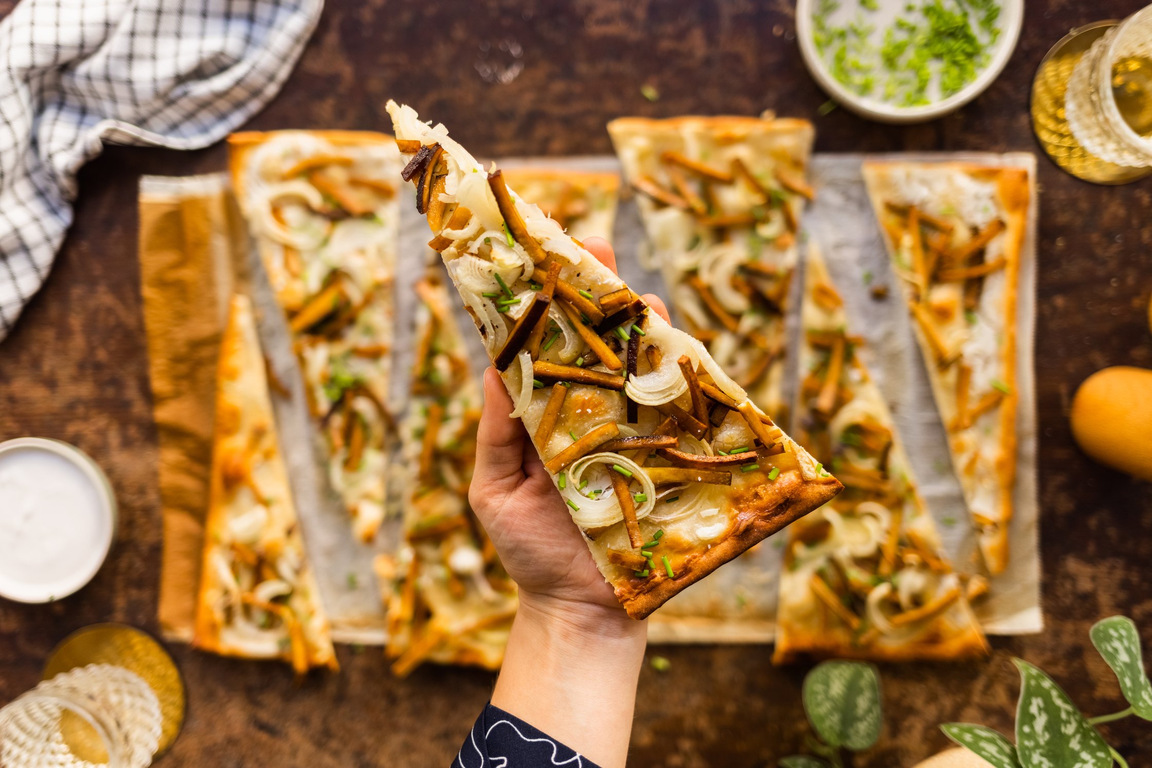 Ein Stück veganer Flammkuchen wird mit Hand hochgehalten, Glas Wasser, Schale mit Creme Fraiche