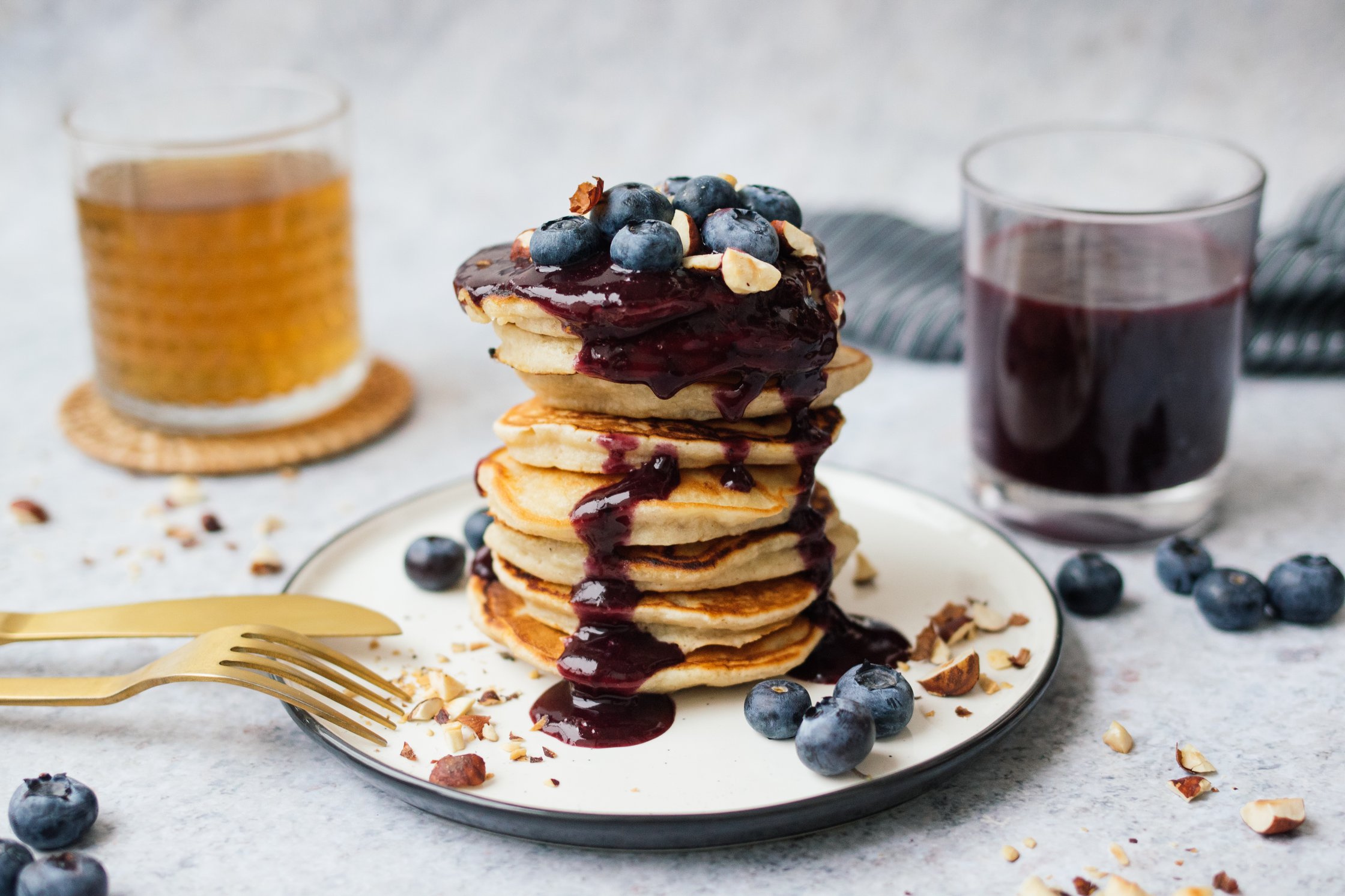 Pancake Stapel auf Teller mit Blaubersoße und Topping aus frischen Blaubeeren und gehackten Haselnüssen, ein Glas Tee und ein