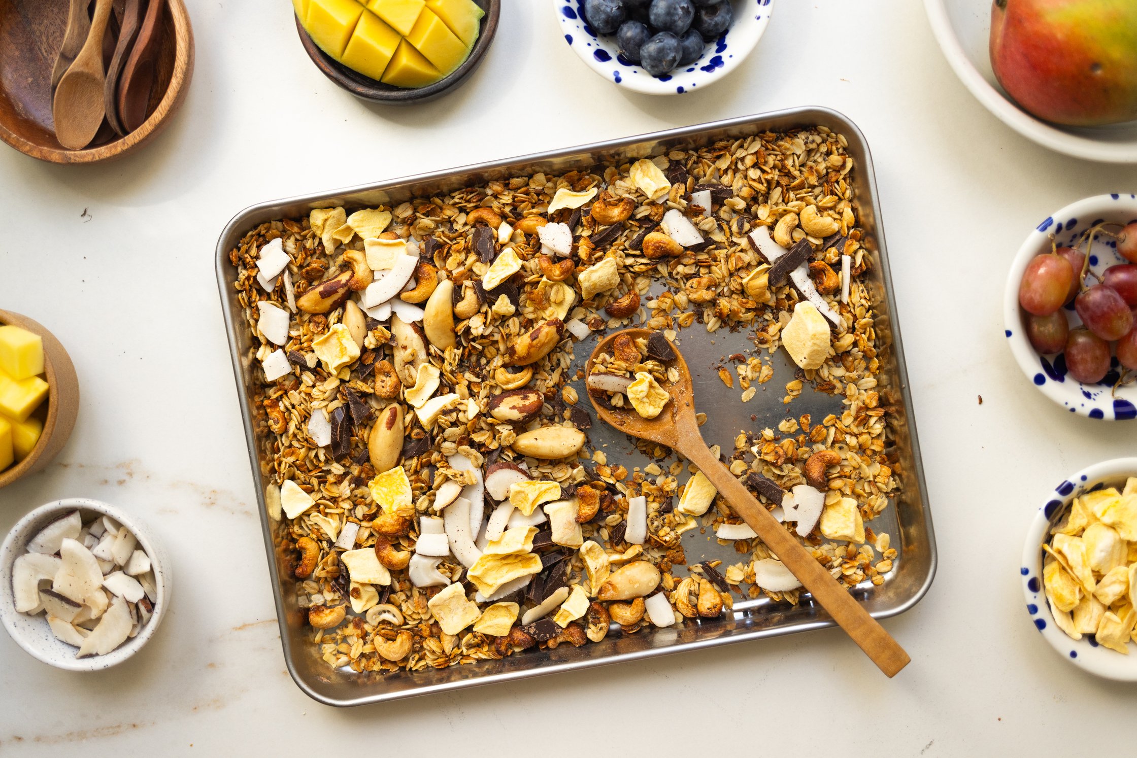 The photo shows a baking sheet filled with granola, viewed from above. Ingredients such as mango, blueberries, pecans, and co