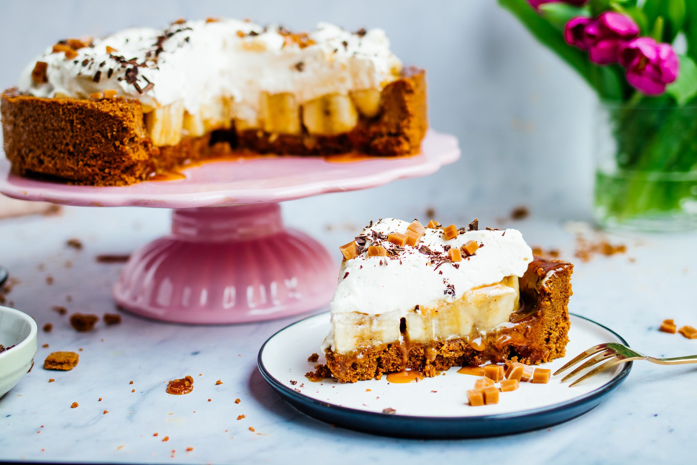 Cake platter with vegan banoffee pie, piece of cake on a plate, hand holding fork with vegan whipped cream and homemade caram