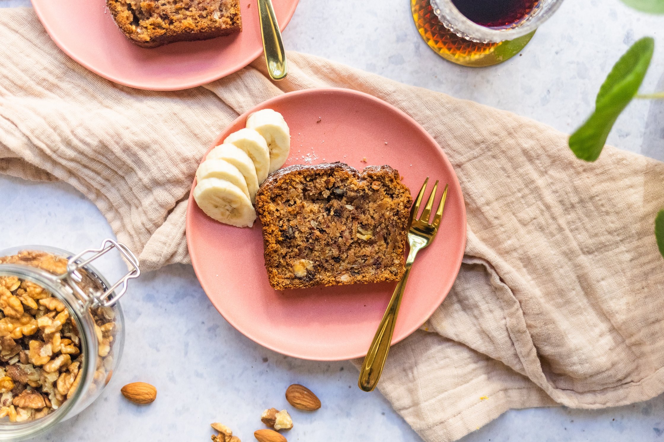 a piece vegan banana bread on a pink plate with banana and a golden fork, a cup of coffee and walnuts in a glas next to it