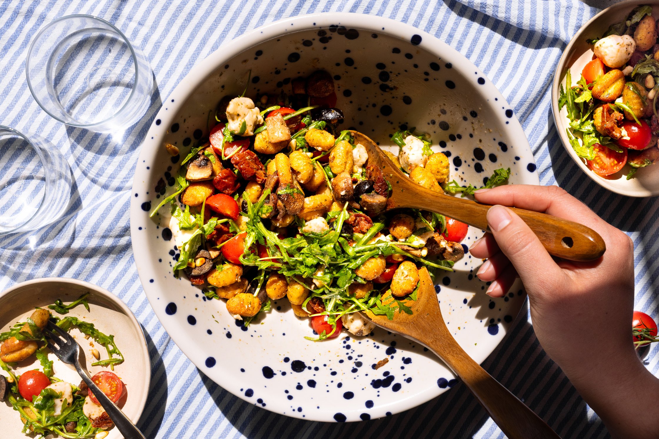 In einer großen weißen Salatschüssel mit blauen Punkten wird der Gnocchi-Salat mit zwei Holzlöffeln vermengt.