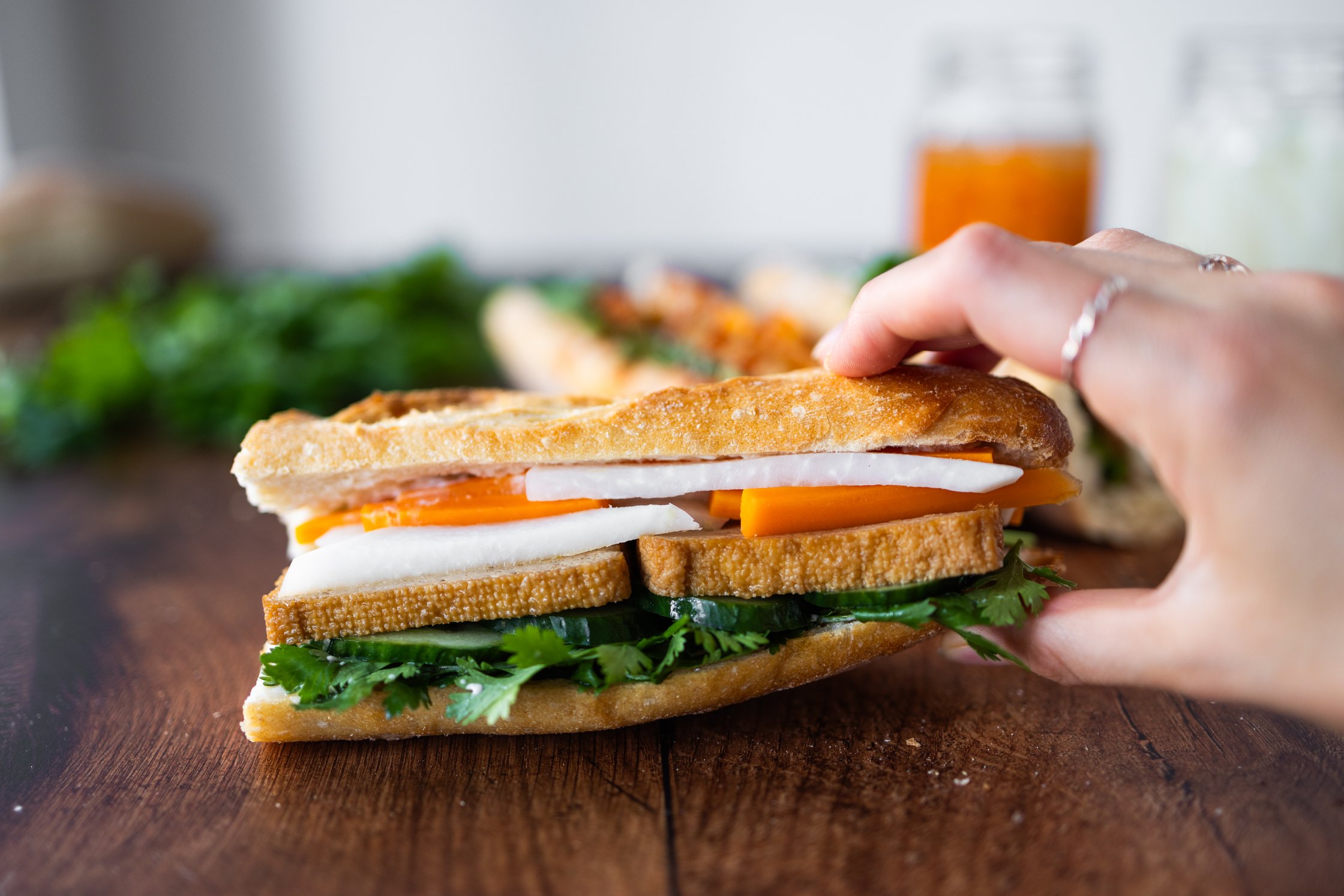 vegan banh mi topped with sriracha, filled with pickled vegetables, tofu, cilantro, vegan mayonnaise, vegan butter, and cucum