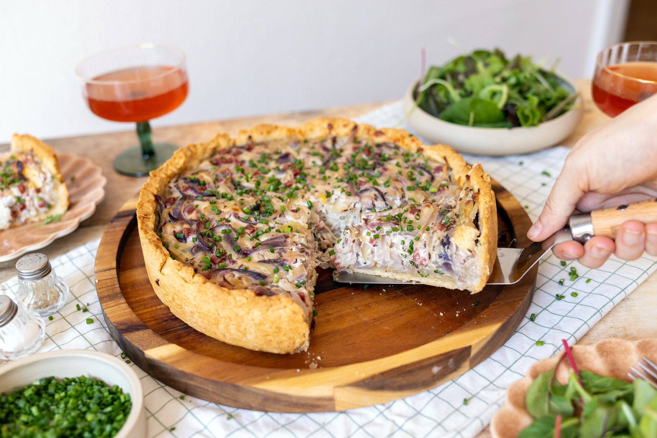 Ein gedeckter Tisch. Der Zwiebelkuchen steht in der Mitte und ein Stück wird rausgehoben.