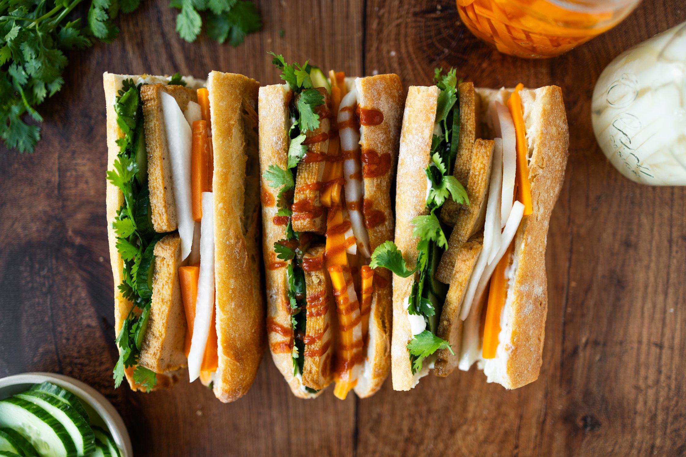 vegan banh mi topped with sriracha, filled with pickled vegetables, tofu, cilantro, vegan mayonnaise, vegan butter, and cucum