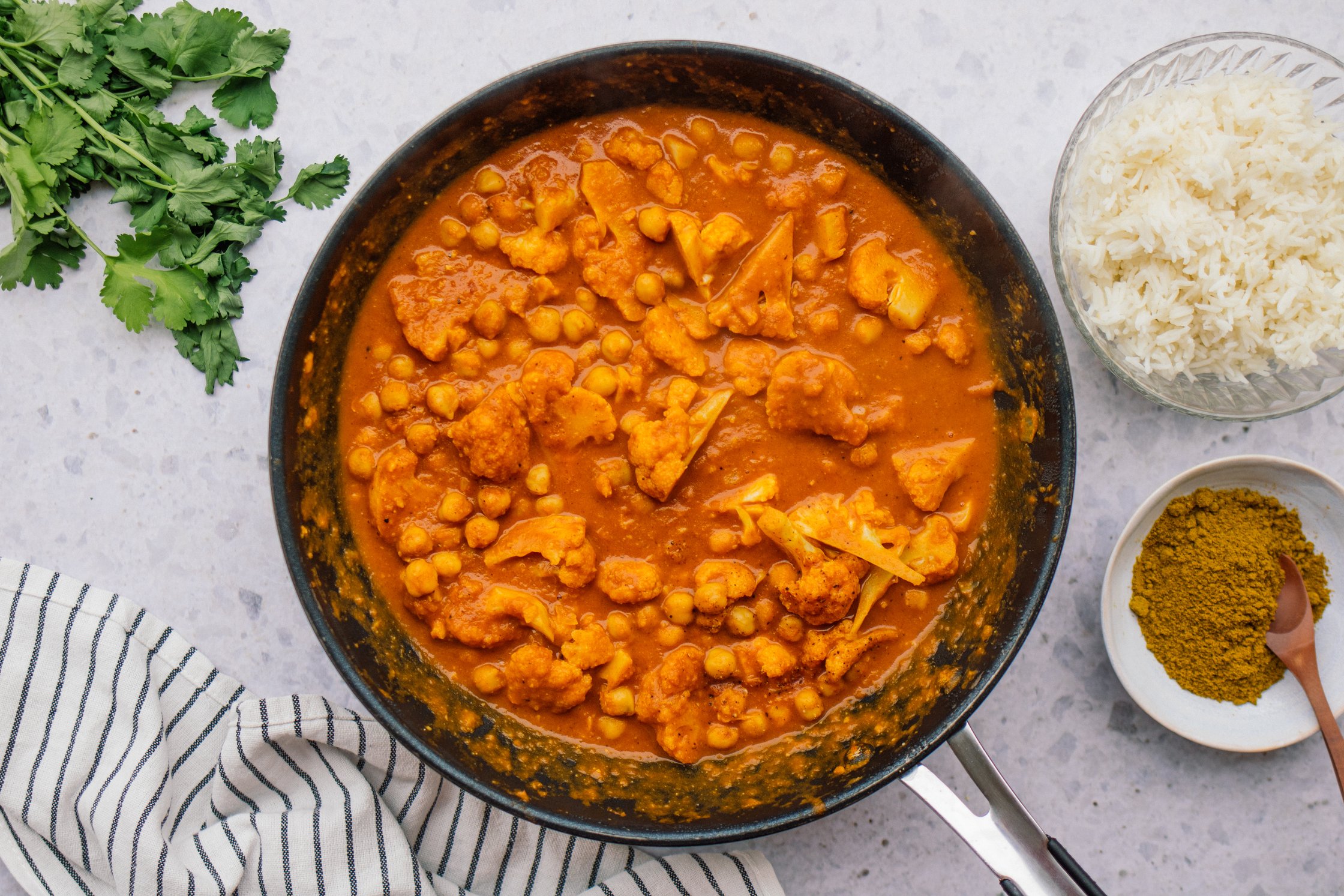 A pan with cooked vegan chicken madras including cauliflower and chickpeas, also bowls of cooked rice and madras curry powder