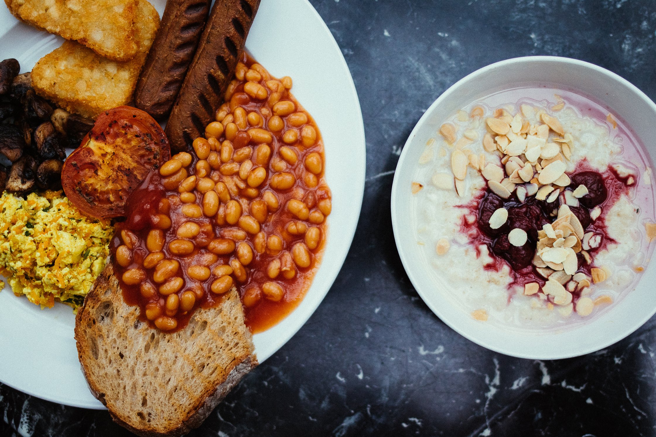 ein Teller mit English Breakfast und eine kleine Schüssel mit Porridge stehen auf einem dunklen Tisch