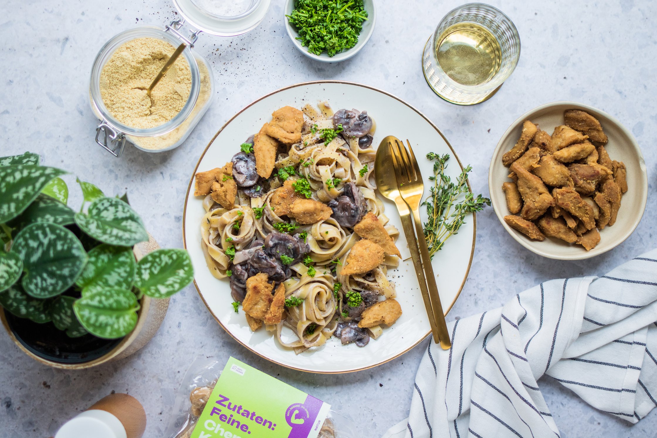 Plate with vegan Chicken, Pasta and Mushrooms, a Glas of Water, a small Bowl with Parsley, Cutlery