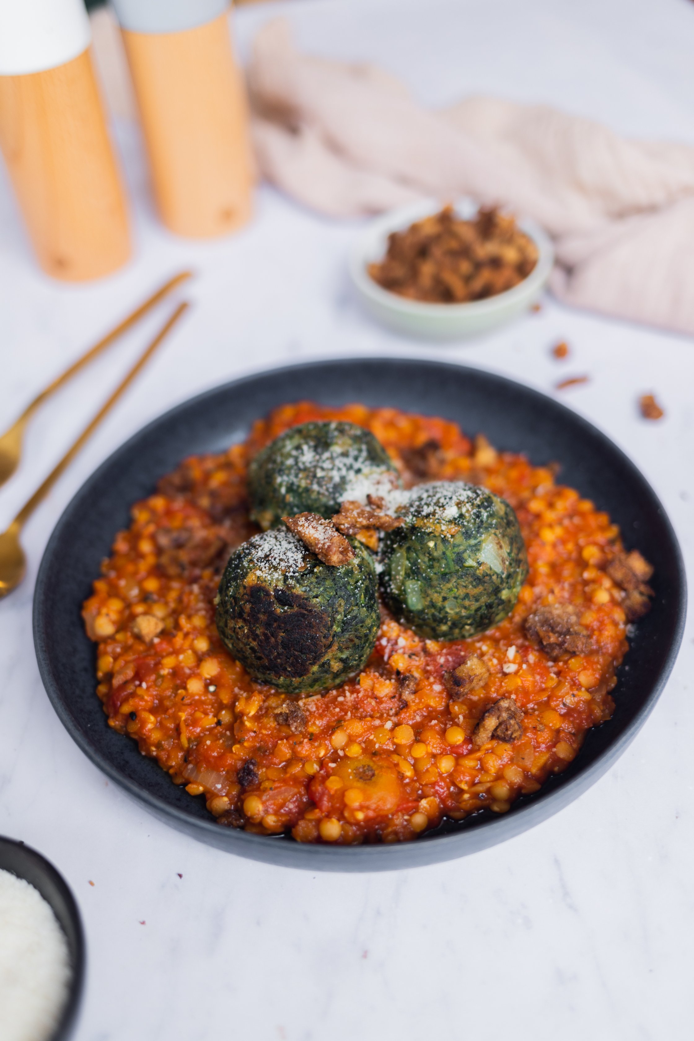 Tomaten-Linsen-Soße und Spinatknödel auf einem Teller mit Zimt-Croutons, Besteck, Salz- und Pfeffermühle daneben