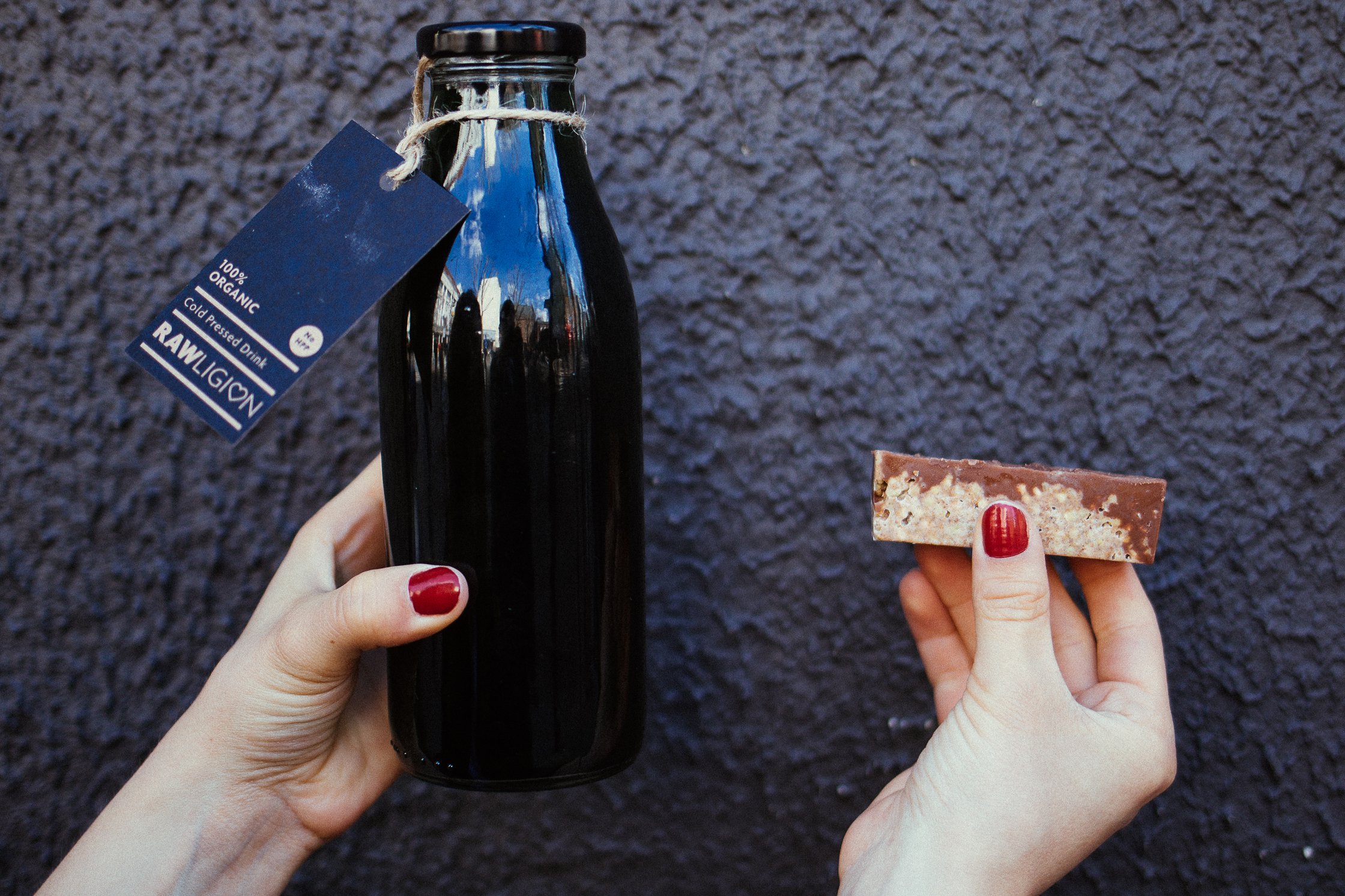 zwei Hände vor grauem Hintergrund, die eine hält eine schwarze Flasche, die andere einen Schokoriegel