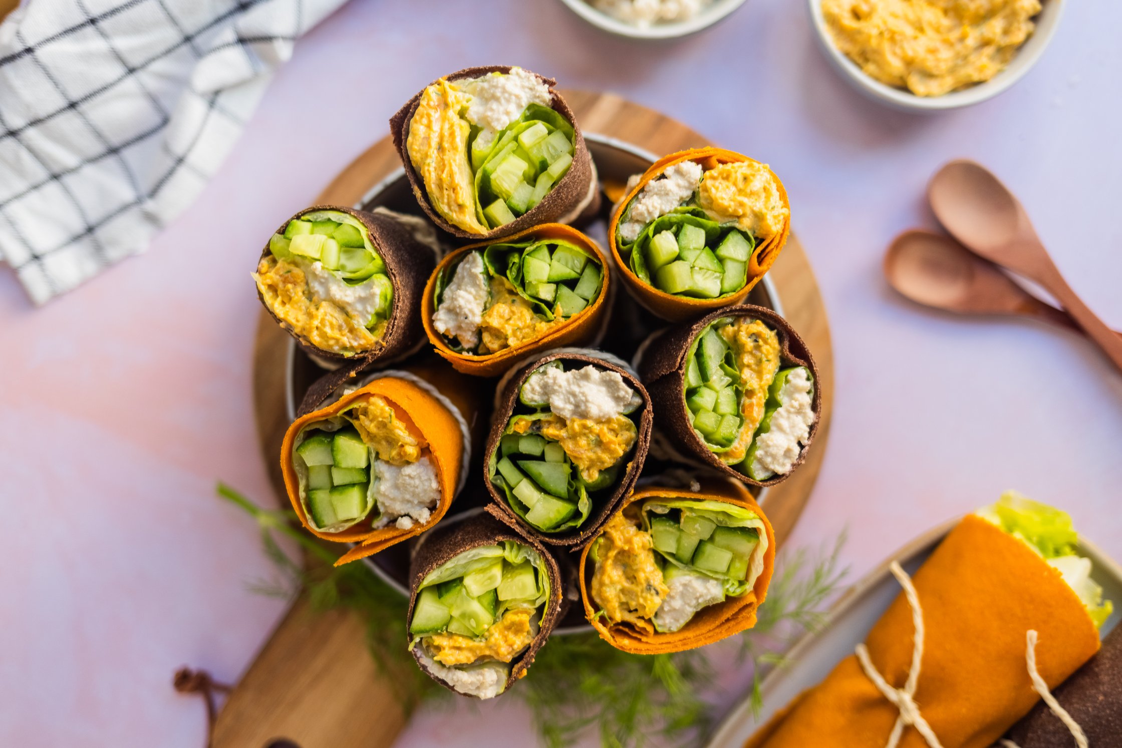 Vegane Gemüse-Wraps in einer Schale auf einem Brettchen, mit einem Küchentuch, zwei Holzlöffeln und Wraps auf einem Teller
